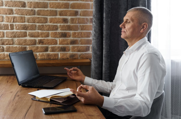 一名商人闭着眼睛坐在家中的工作场所，一边在笔记本电脑上工作，一边冥想。图片下载