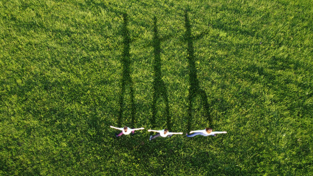 三名年轻女子在日落时分的草地上练习瑜伽。从上面看。草地上长长的影子图片下载