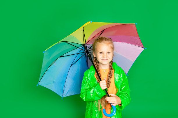 一个小女孩在一个孤立的绿色背景，一把伞和一件雨衣微笑，文字的空间图片下载