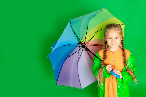 一个小女孩在一个孤立的绿色背景，一把伞和一件雨衣微笑，文字的空间图片下载