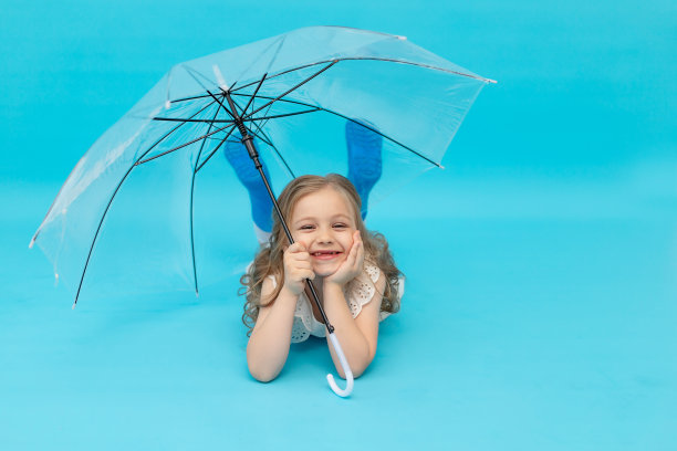 一个快乐可爱的小女孩穿着蓝色胶靴和纯棉白色连衣裙撑着伞躺在蓝色背景的工作室里笑着，笑着，嬉闹着，一个文字的地方图片下载