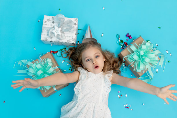 可爱的惊喜小女孩在棉质白色连衣裙上的蓝色背景，在节日的帽子与礼物和五彩纸屑，文字的地方图片下载
