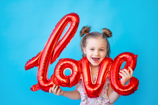 一个小女孩在一个蓝色的孤立的工作室背景上，红色的气球在题词“爱”上玩得很开心。用于复制文本的空间。情人节概念图片下载