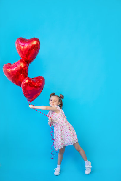一个小女孩在蓝色的孤立工作室背景上，红色的气球在一个心形的地方玩得很开心。用于复制文本的空间。情人节概念图片下载