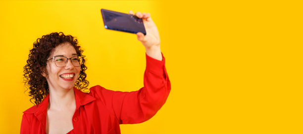 带着眼镜的漂亮卷发女孩在自拍。一位年轻女子用手机给自己拍照。工作室女性肖像在明亮的黄色背景。图片下载