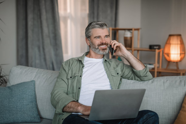 开朗的欧洲老人与胡子打电话给客户和笔记本电脑在客厅内部工作图片下载