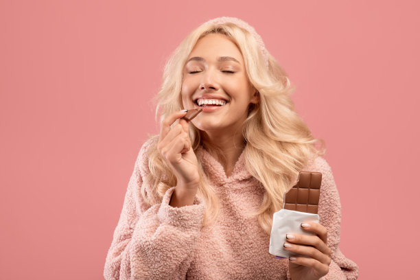 年轻的白人女士吃巧克力，享受美味的甜点闭着眼睛，站在粉红色的工作室背景图片下载