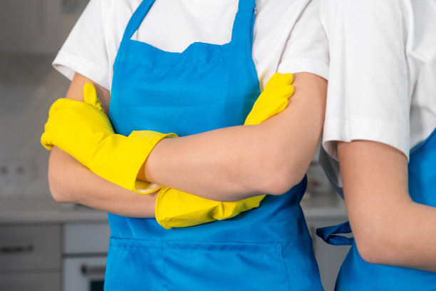 两个人的清洁团队，他们的手臂交叉在胸前，背景是一个模糊的厨房。专业女工戴着围裙和黄色橡胶手套清洁公司。图片下载