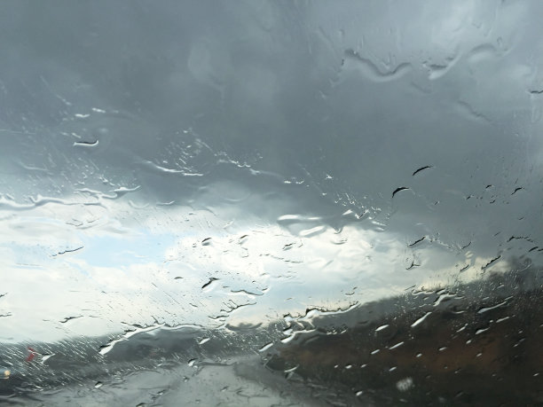 下雨天开车。雨点落在汽车挡风玻璃上，多云的天空和空旷的道路背景图片下载