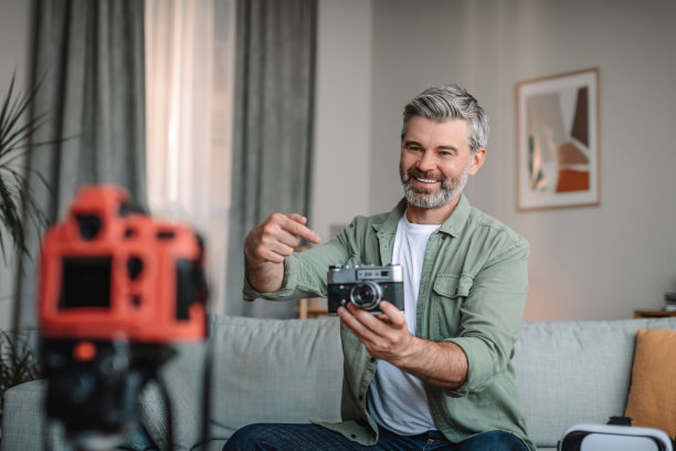 在家写博客。快乐的白种人大胡子男子展示照片相机和拍摄视频在房间里图片下载