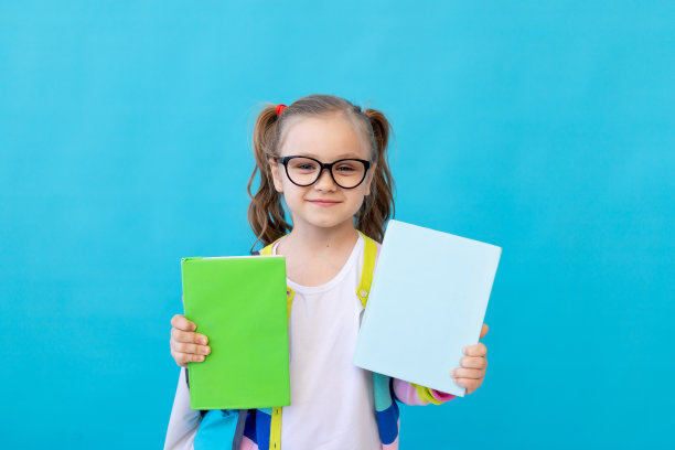 一个可爱的小女孩的肖像，戴着眼镜，穿着条纹夹克，有两本空书。教育理念。照相馆，蓝色背景，文字的地方图片下载