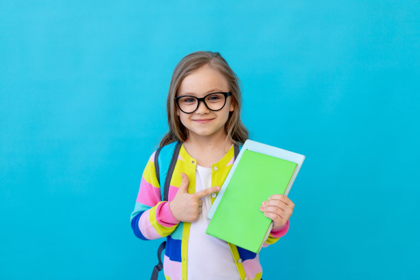 可爱的女孩孩子女学生眼镜在一个蓝色孤立的背景，公文包和书在她的手显示类和微笑，文字空间图片下载