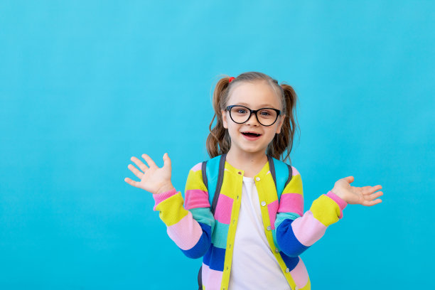 一个可爱的小女孩的肖像，戴着眼镜，穿着条纹夹克，手里拿着笔记本，背着背包，高兴和快乐。教育理念。蓝色背景，文本的位置图片下载