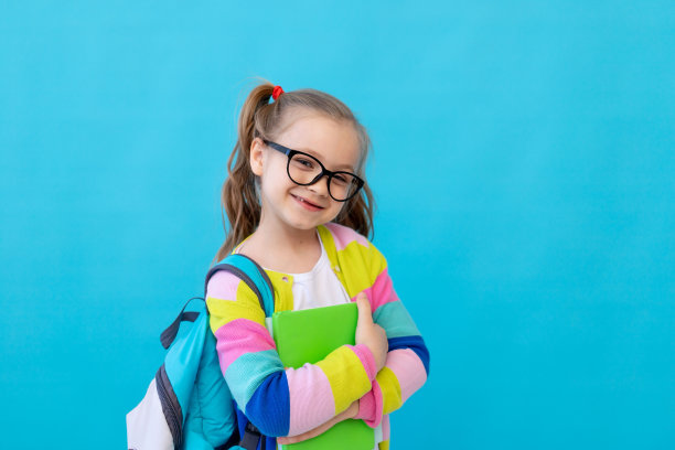 一个可爱的小女孩的肖像，戴着眼镜，穿着条纹夹克，手里拿着笔记本和教科书，背着背包微笑着。教育理念。照相馆，蓝色背景，文字的地方图片下载