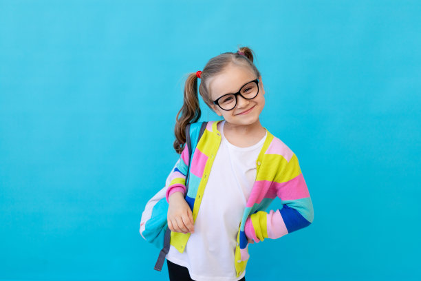一个可爱的小女孩的肖像，戴着眼镜，穿着条纹夹克和背包。教育理念。照相馆，蓝色背景，文字的地方图片下载