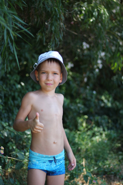 一名身穿泳裤、头戴帽子的欧洲白人男生在热带树叶的背景下竖起大拇指。凉爽的暑假。本空间图片下载