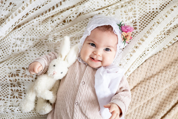一个穿着自然米色衣服的小女孩躺在家里的床上，手里拿着一只柔软的玩具兔子微笑着，特写照片图片下载