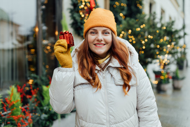 冬天，一名妇女在室外拿着一份圣诞礼物。降雪。冷杉树，后面有花环。购物的概念，节日和快乐的圣诞节。图片下载