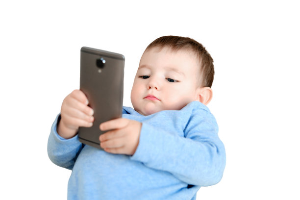 一个蹒跚学步的小男孩带着手机坐在客厅的沙发上，孤立地坐在白色背景上。孩子在床上，手里拿着智能手机图片下载