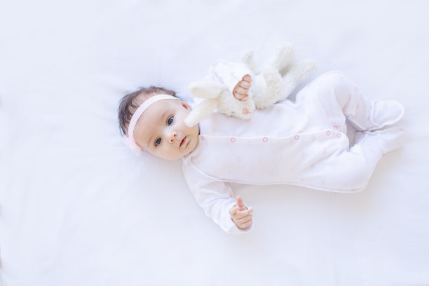 女婴微笑着躺在白色棉床上，带着玩具入睡或早上醒来，可爱的新生小婴儿在家里一个孤立的背景特写图片下载