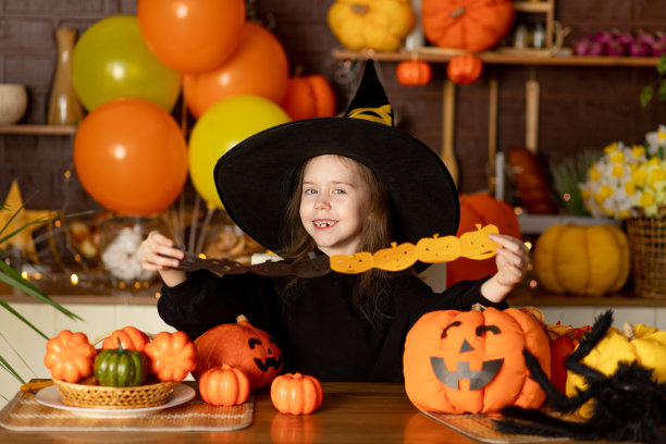 万圣节前夕，一个穿着女巫服装、带着南瓜和大蜘蛛的小女孩在黑暗的厨房里用剪刀剪出节日的花环，微笑着，高兴着图片下载
