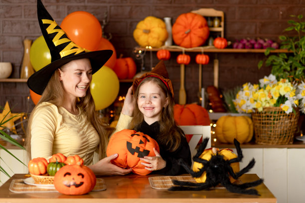 万圣节前夕，一个孩子的母亲和女儿穿着女巫服装，有南瓜和一只大蜘蛛在黑暗的厨房里微笑和欢乐地庆祝万圣节假期图片下载