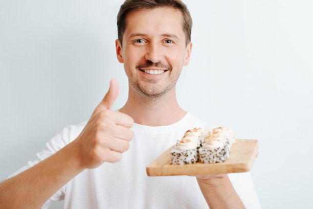 快乐的英俊的年轻男子的肖像，显示喜欢的姿态，拿着寿司在木板室内。微笑的家伙喜欢亚洲传统美食看着相机。图片下载