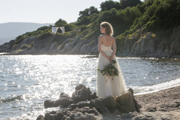 新娘穿着长长的白色婚纱站在风多的海岩岸边，手里拿着郁郁葱葱的绿色波西米亚花束图片下载