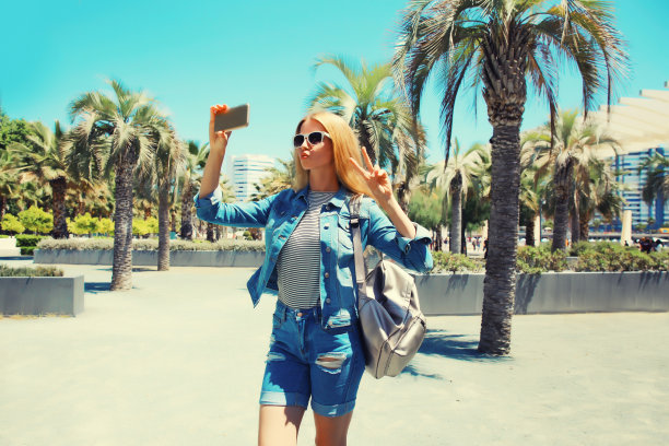 年轻女子在夏天公园用手机自拍的肖像，背着背包，背景是棕榈树图片下载