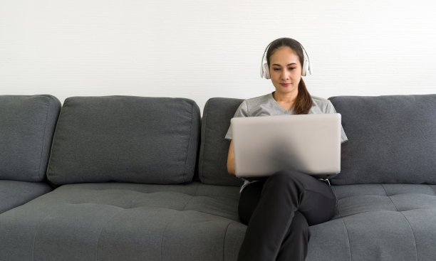穿着灰色t恤的亚洲中年妇女在客厅用笔记本电脑打字，一边听耳机音乐，享受悠闲的周末。无压力概念图片下载