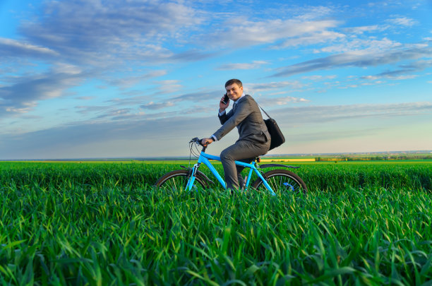 一个商人骑着自行车在绿油油的草地上，穿着西装，他在打一个电话，美丽的大自然在春天，自由职业者的商业概念图片下载