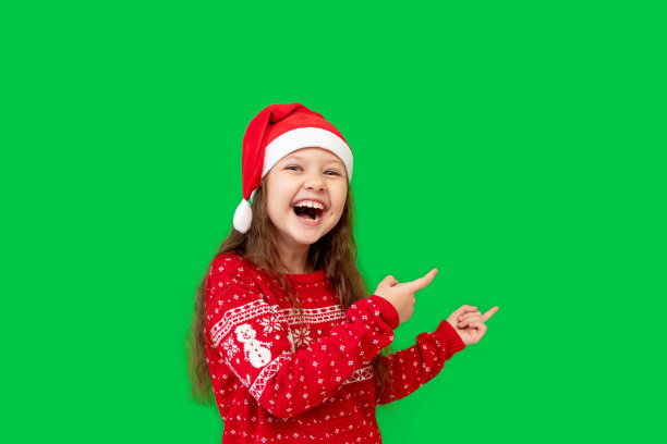 一个快乐的小女孩穿着冬装，戴着圣诞老人的帽子，红色的毛衣在绿色的孤立背景上，她伸出手指，微笑着，一个地方或空间的文字图片下载