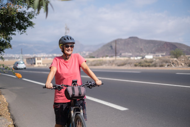 笑容满面的老年妇女，戴着防护头盔，骑着她的电动自行车在阳光明媚的道路上奔跑。健康的生活方式和可持续的出行理念图片下载