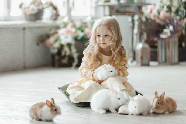 一个穿着黄色连衣裙的微笑的小女孩在一间屋子的地板上和兔子玩耍。一个婴儿和一只兔子。客厅为复活节而装饰。图片下载