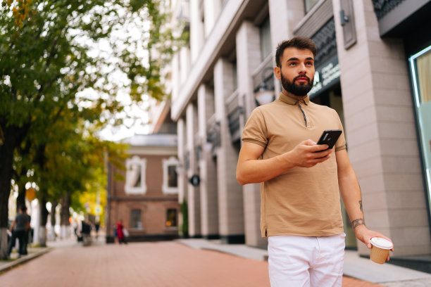 低角视角，夏日里，手有纹身的自信年轻人走在城市街道上，用智能手机喝着外卖咖啡。图片下载