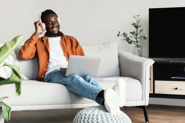 兴奋的非裔美国人戴着耳机使用笔记本电脑，坐在家里的沙发上在线工作，自由空间图片下载