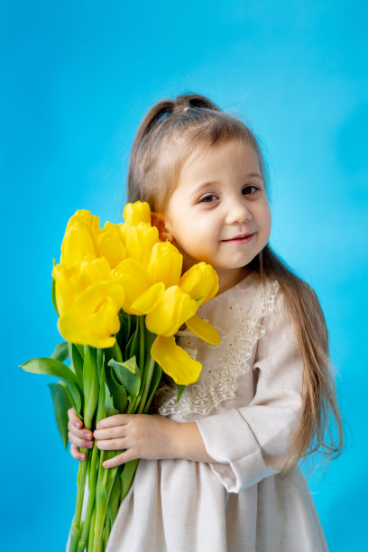 一个微笑的小女孩的肖像，一个孩子与一束黄色郁金香在蓝色孤立的背景。的生活方式。国际妇女节或母亲节。文本空格。高质量的摄影图片下载