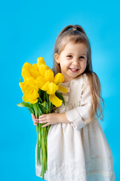 一个微笑的小女孩的肖像，一个孩子与一束黄色郁金香在蓝色孤立的背景。的生活方式。国际妇女节或母亲节。文本空格。高质量的摄影图片下载