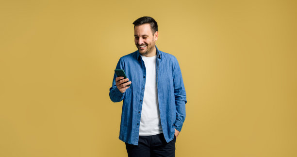 微笑中成人英俊男子浏览社交媒体上的智能手机，而摆姿势与手在口袋上孤立的黄色背景图片下载