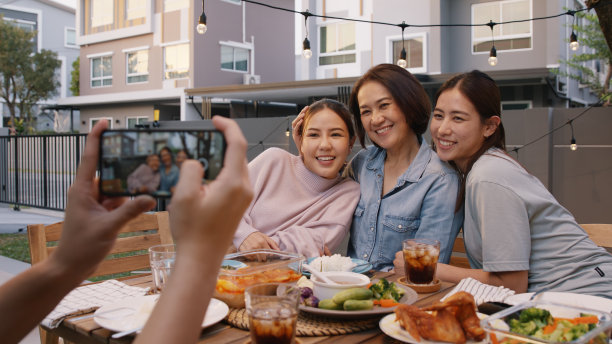年轻的成年人亚洲人拥抱拥抱亲吻爱照顾妈妈拍照自拍视频在手机上相机在家里野餐吃饭有趣的晚上聚会餐桌。放松年长的妈妈，微笑着享受温暖的时光。图片下载