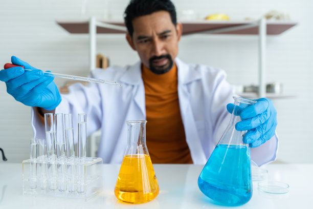 一位穿着白色白大褂、戴着橡胶手套的男老师，架子上和桌子上放着许多实验工具。右手拿移液管，左手拿蓝色化学瓶。在化学课开始之前。图片下载