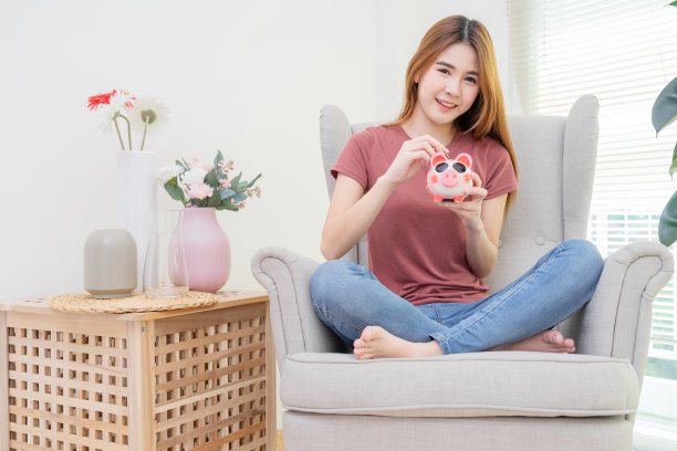 年轻女子非常高兴地把硬币放进存钱罐，放在靠近窗户和花瓶的沙发上图片下载