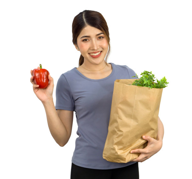 年轻的亚洲女子穿着便服微笑着站在那里，手里拿着一个红色的甜椒，另一只手拿着一个装满蔬菜的纸袋。人像在白色背景与工作室灯光。孤立的图片下载