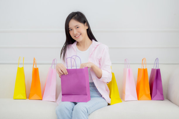 幸福年轻的亚洲妇女坐在沙发上打开纸袋，而网上购物与信用卡在家里，女孩购买和折扣的消费者兴奋，购物狂和生活方式的概念。图片下载