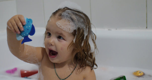 在泡沫浴中沐浴的女孩脸上稚气的表情被升华了。小女孩看着泡沫里的玩具惊讶地尖叫起来。孩子把玩具从泡沫中拉了出来。图片下载