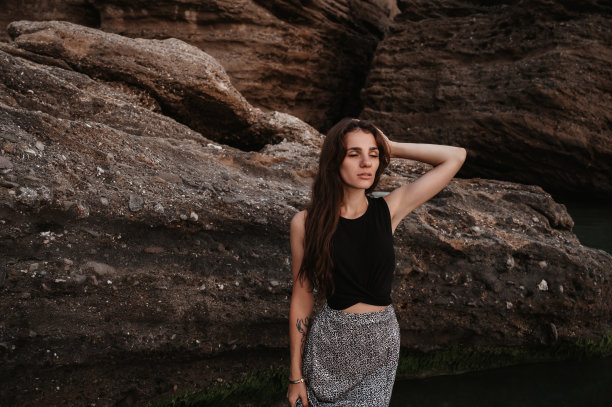 一张海滩照片，一个美丽的性感的年轻女子，黑色长发，穿着裙子，在海滩上的巨石旁摆姿势。图片下载