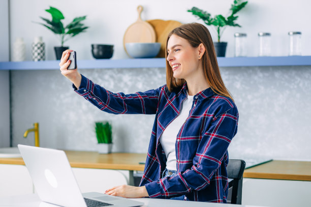 快乐有魅力的女人坐在家里的厨房里，使用智能手机与家人通话，朋友告诉好消息，感觉积极，自拍。图片下载