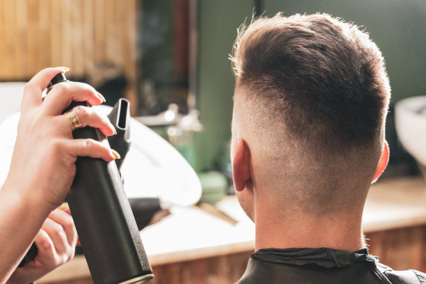 在理发店里，一名女理发师用吹风机向坐在椅子上的年轻男子喷洒喷雾并吹干头发图片下载