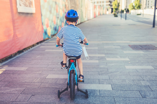 在新冠肺炎疫情期间，可爱的小男孩戴着防护口罩和安全帽在阳光明媚的夏日在公园骑自行车，新常态的生活方式概念。图片下载