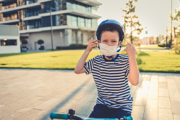 新常态蹒跚学步的男孩戴着防护面具和头盔骑自行车。一个男孩一边骑自行车一边调整他脸上的面具。图片下载
